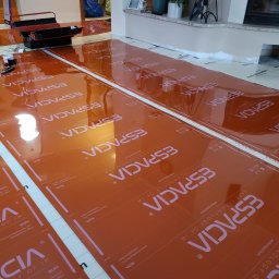 ESPACIA Sp. z o.o. - Pomarańczowe maty grzewcze marki Espacia rozłożone na podłodze w trakcie montażu, widoczne narzędzia i akcesoria montażowe, w tle kominek i elementy wystroju wnętrza.