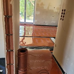 ESPACIA Sp. z o.o. - Pomarańczowe maty grzewcze marki Espacia rozłożone na czarnej folii, widoczne przewody i zwinięta siatka montażowa, surowy stan ścian i podłogi w trakcie instalacji.