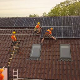 ESPACIA Sp. z o.o. - Montaż paneli fotowoltaicznych na dachu z brązowej dachówki; czterech monterów w pomarańczowych koszulkach i żółtych kaskach zabezpieczonych linami, pracuje nad instalacją paneli słonecznych...