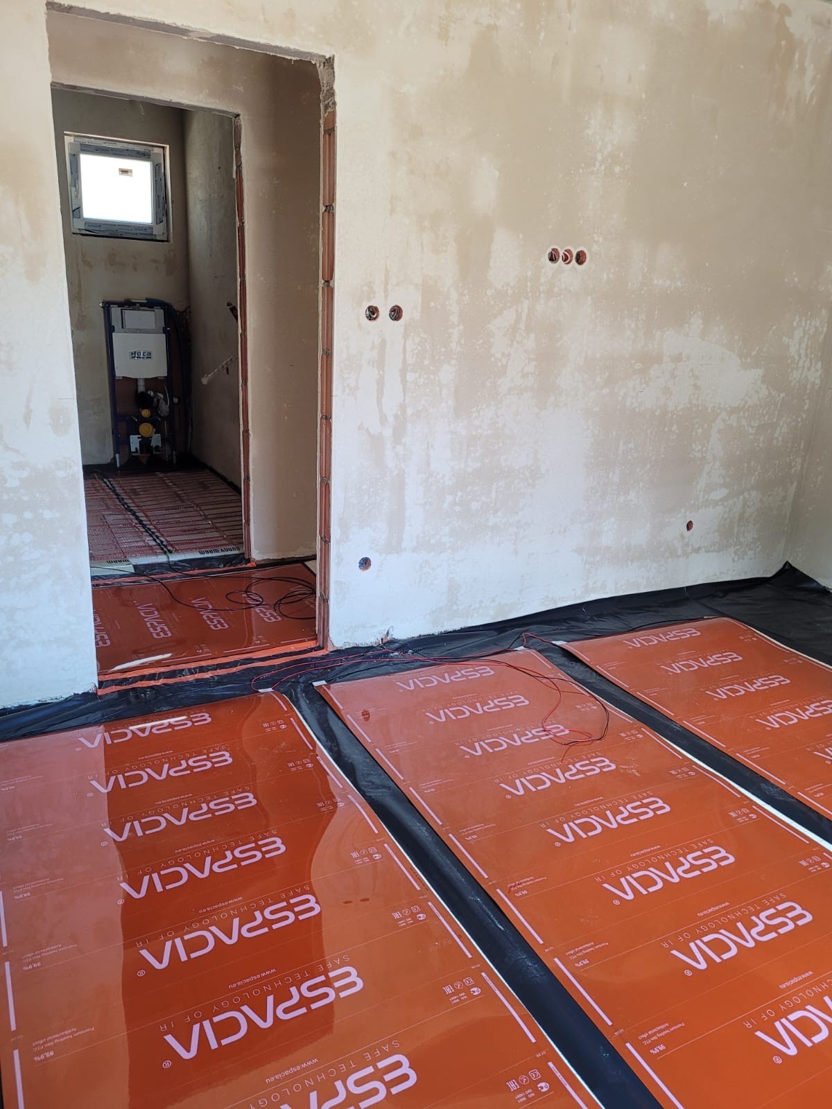 Montaż mat grzewczych Espacia na czarnej folii w surowym pomieszczeniu z widoczną instalacją sanitarną w tle.