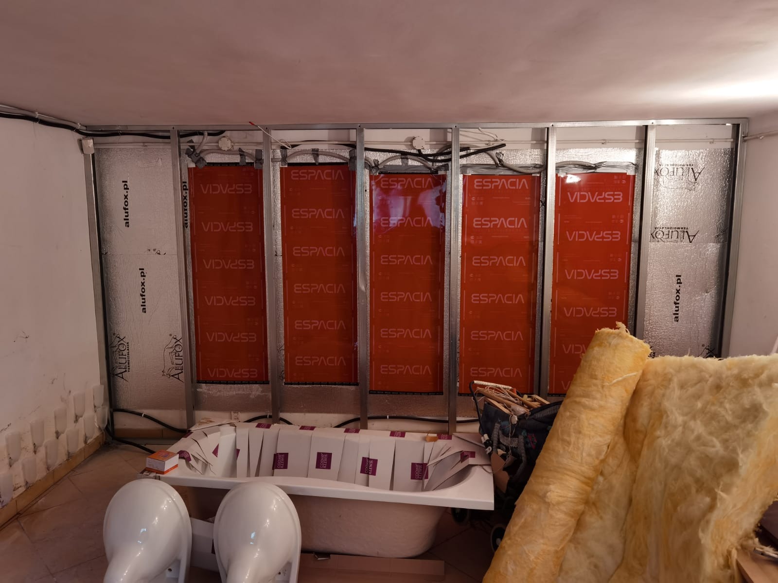 Montaż paneli grzewczych Espacia na ścianie, widoczna instalacja stelaża, izolacja z wełny mineralnej oraz elementy wyposażenia łazienki: wanna i dwie umywalki.