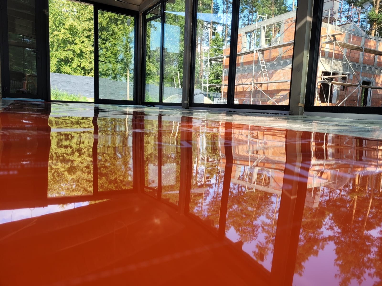Błyszcząca, pomarańczowa posadzka żywiczna w nowym budynku z dużymi oknami, odbijająca widok drzew i budowy w tle.