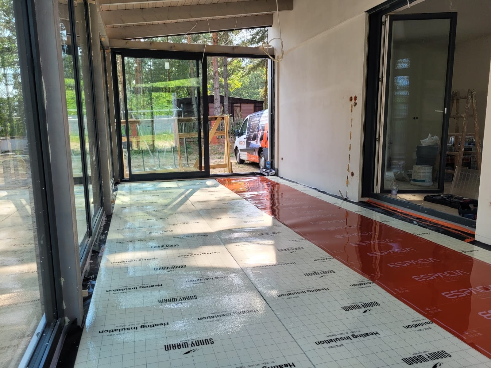 Instalacja ogrzewania podłogowego: widok na rozłożone maty grzewcze 'Sunny Warm' i 'Espacia' na podkładzie izolacyjnym w przeszklonym pomieszczeniu z widokiem na zewnątrz, częściowo pokryte...