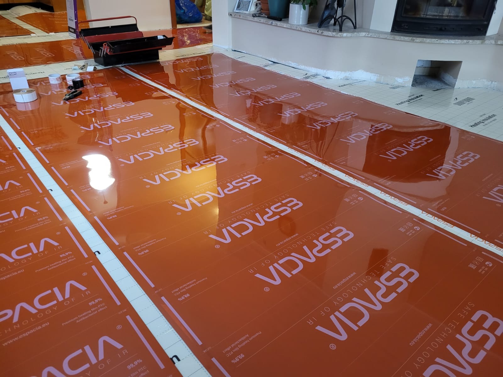 Pomarańczowe maty grzewcze marki Espacia rozłożone na podłodze w trakcie montażu, widoczne narzędzia i akcesoria montażowe, w tle kominek i elementy wystroju wnętrza.