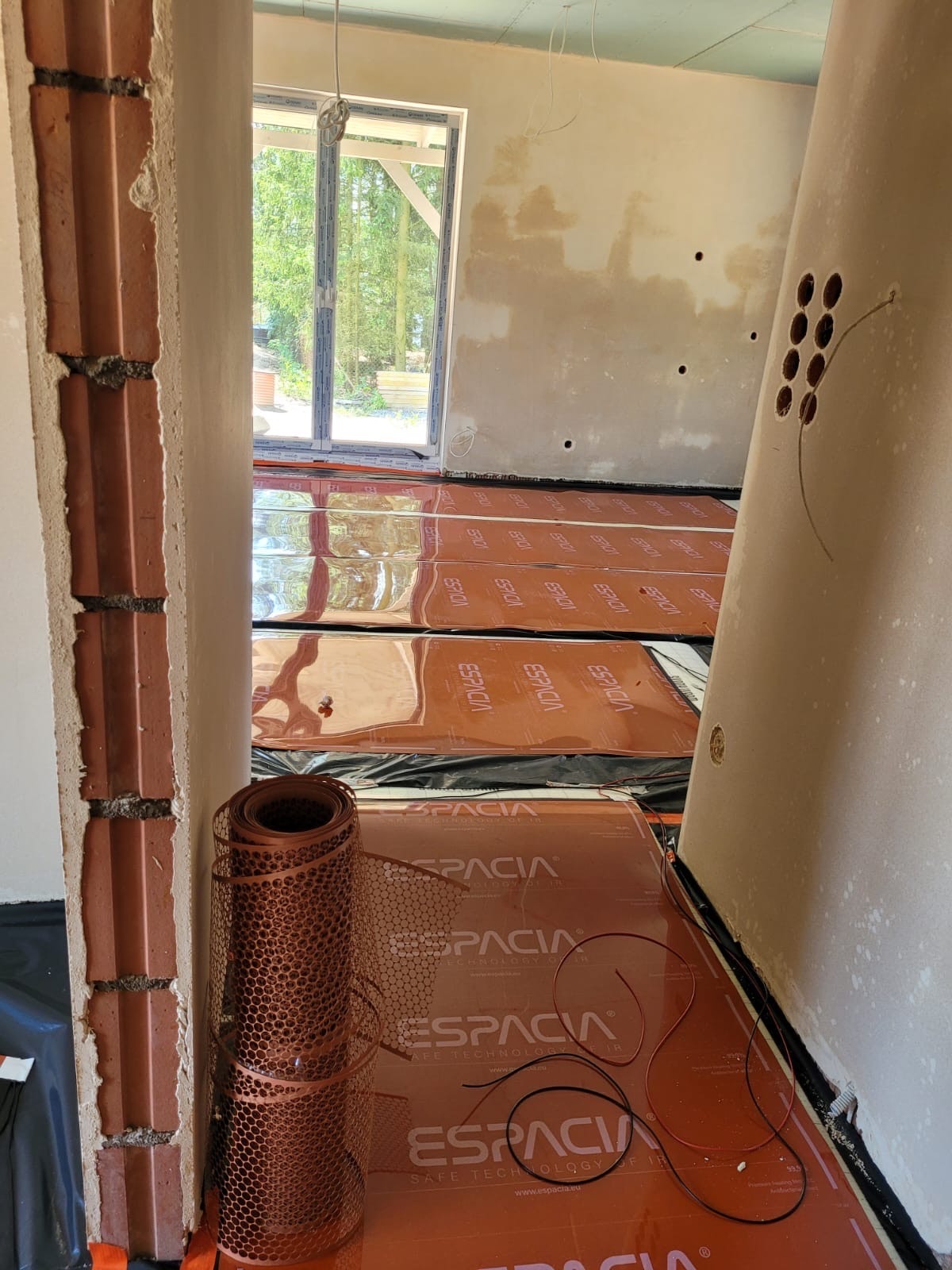 Pomarańczowe maty grzewcze marki Espacia rozłożone na czarnej folii, widoczne przewody i zwinięta siatka montażowa, surowy stan ścian i podłogi w trakcie instalacji.