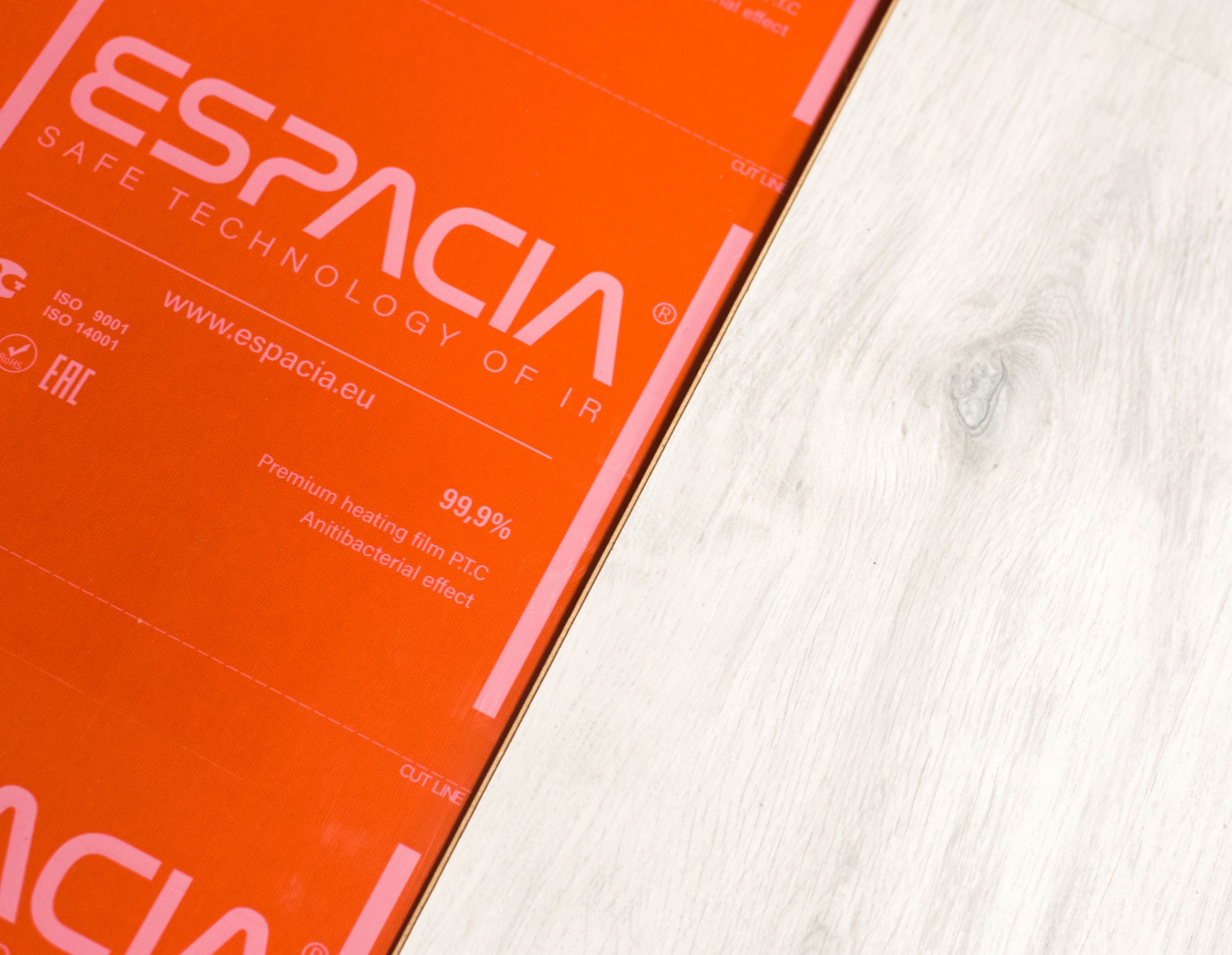 Pomarańczowa mata grzewcza marki Espacia z widocznym logo i informacjami o produkcie, ułożona obok jasnej drewnianej podłogi.