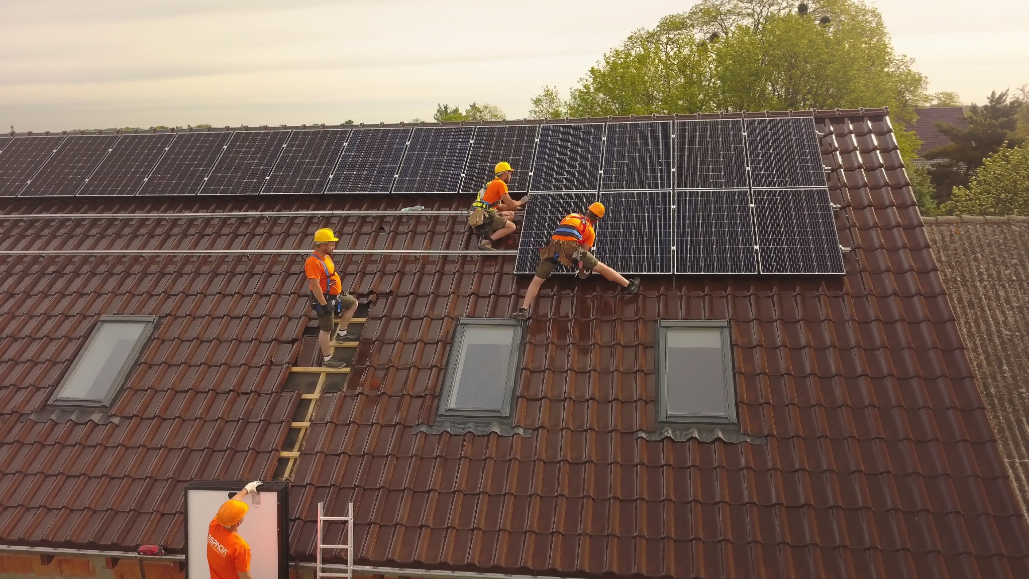 Montaż paneli fotowoltaicznych na dachu z brązowej dachówki; czterech monterów w pomarańczowych koszulkach i żółtych kaskach zabezpieczonych linami, pracuje nad instalacją paneli słonecznych...