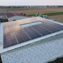 Instalacja paneli słonecznych na dachu budynku gospodarczego z białej cegły, widok z góry na wiejski krajobraz.