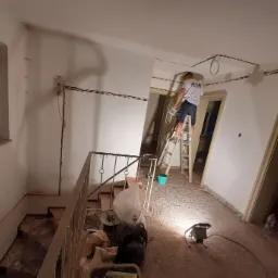 Instalacja przewodów elektrycznych w korytarzu: mężczyzna na drabinie montuje kable pod sufitem, widoczne przewody poprowadzone wzdłuż ścian, surowy stan wykończenia, podłoga pokryta kurzem...