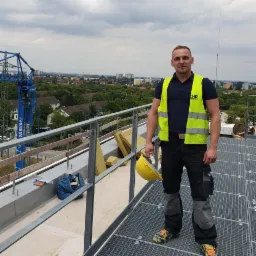 Elektryk w żółtej kamizelce i kasku na dachu budynku z widokiem na miasto, na tle błękitnego żurawia budowlanego.