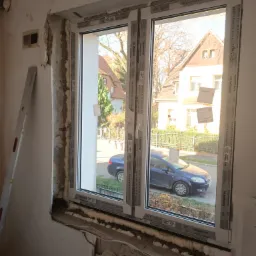 Montaż okna PCV z widoczną pianką montażową i uszkodzonym tynkiem, na zewnątrz widok na ulicę z domami i zaparkowanym niebieskim samochodem.