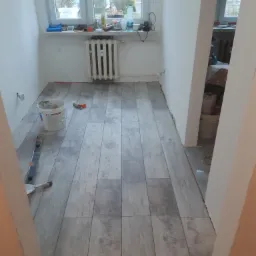 Ułożone panele podłogowe imitujące drewno w trakcie remontu pokoju, widoczny poziomica, wiadro z klejem i szpachelka, kaloryfer pod oknem.