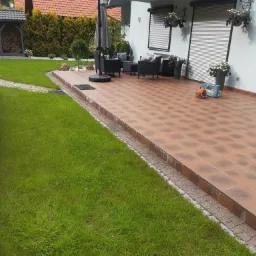 Brązowa, kwadratowa płytka tarasowa z obrzeżem z kostki, meble ogrodowe z parasolem, skrzynki z kwiatami na ścianie domu, trawnik i drewutnia w tle.