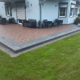 Wykończony taras z brązową, wzorzystą nawierzchnią, obrzeżony ciemnoszarą płytką i kostką, z kompletem mebli ogrodowych w tle, widok z poziomu trawnika.