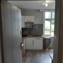 Widok przez otwarte drzwi na odnowioną kuchnię z białymi szafkami, wzorzystą mozaiką nad blatem i jasną podłogą imitującą drewno. Przy oknie stoi walizka w panterkę. W tle widok na zieleń za oknem.