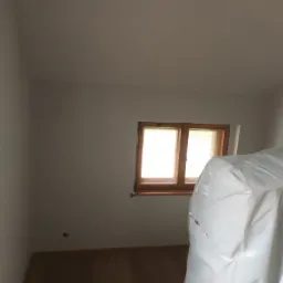 Pomalowane na szaro wnętrze pokoju z drewnianym oknem, podłogą i meblami zabezpieczonymi folią malarską.