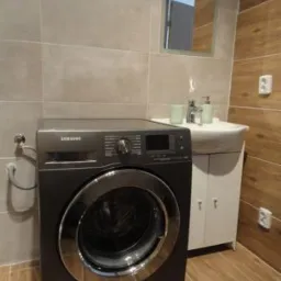 Ciemna pralka Samsung z chromowanym wykończeniem w wyremontowanej łazience z beżowymi i drewnopodobnymi płytkami, obok biała szafka z umywalką i lustrem.