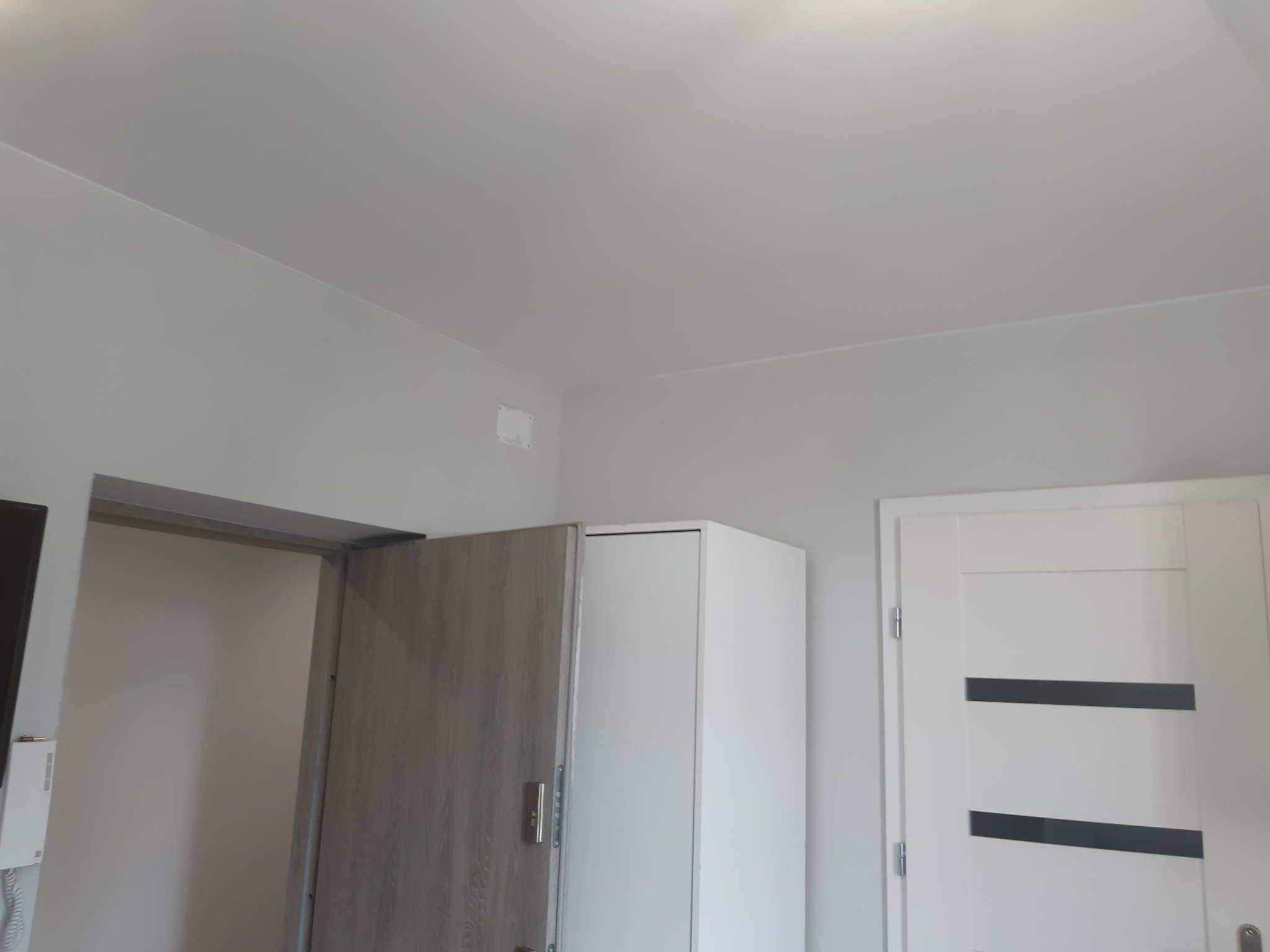 Wnętrze pokoju po odświeżeniu, ściany pomalowane na szaro, widoczna biała szafa i białe drzwi z poziomymi czarnymi paskami.