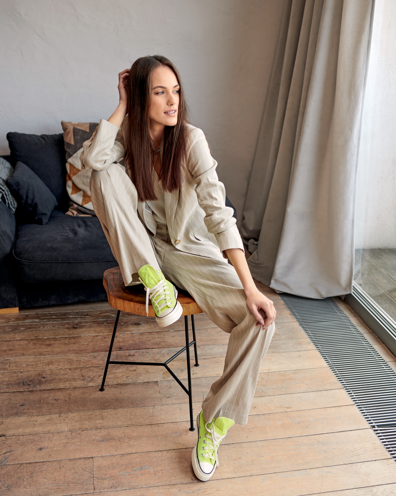 Kobieta w beżowym garniturze w paski i neonowych sneakersach siedzi na drewnianym stołku, spoglądając w bok.
