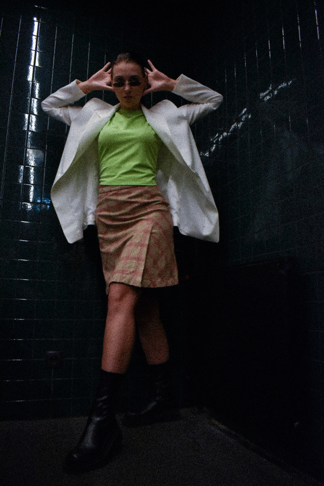 Kobieta w białej marynarce, zielonej bluzce i spódnicy w kratę, pozująca na tle ciemnej ściany z płytek, w czarnych botkach.