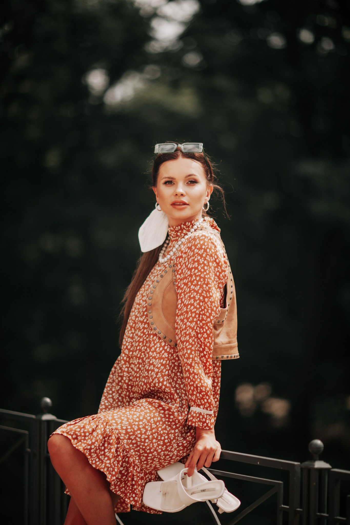 Kobieta w kwiecistej sukience i skórzanej kamizelce, z okularami na głowie, siedzi na metalowym ogrodzeniu, trzymając białe sandały.
