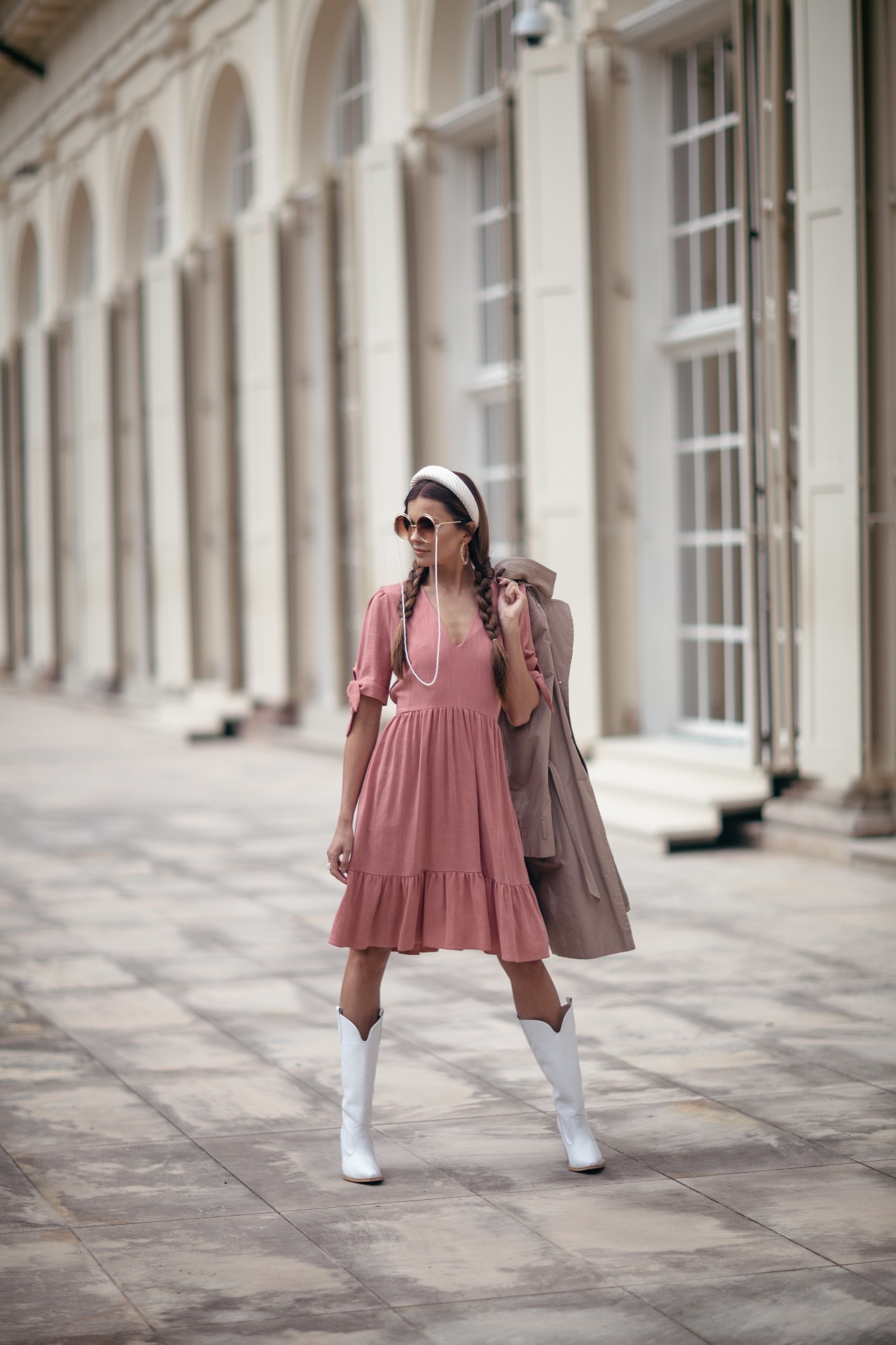 Kobieta w różowej sukience, białych kozakach i okularach na łańcuszku, trzymająca beżowy płaszcz na ramieniu, pozuje na tle kolumnady.