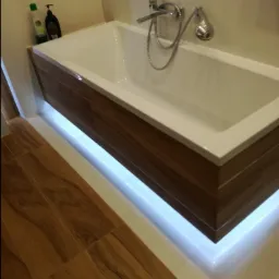 Nowoczesna wanna obudowana drewnopodobnymi panelami z subtelnym podświetleniem LED, bateria chromowana z prysznicem, płytki podłogowe imitujące drewno.