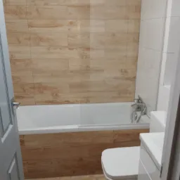 Wykończona łazienka z wanną obudowaną płytkami imitującymi drewno, białą armaturą i częściowo przeszkloną ścianką prysznicową, widok zza uchylonych drzwi.