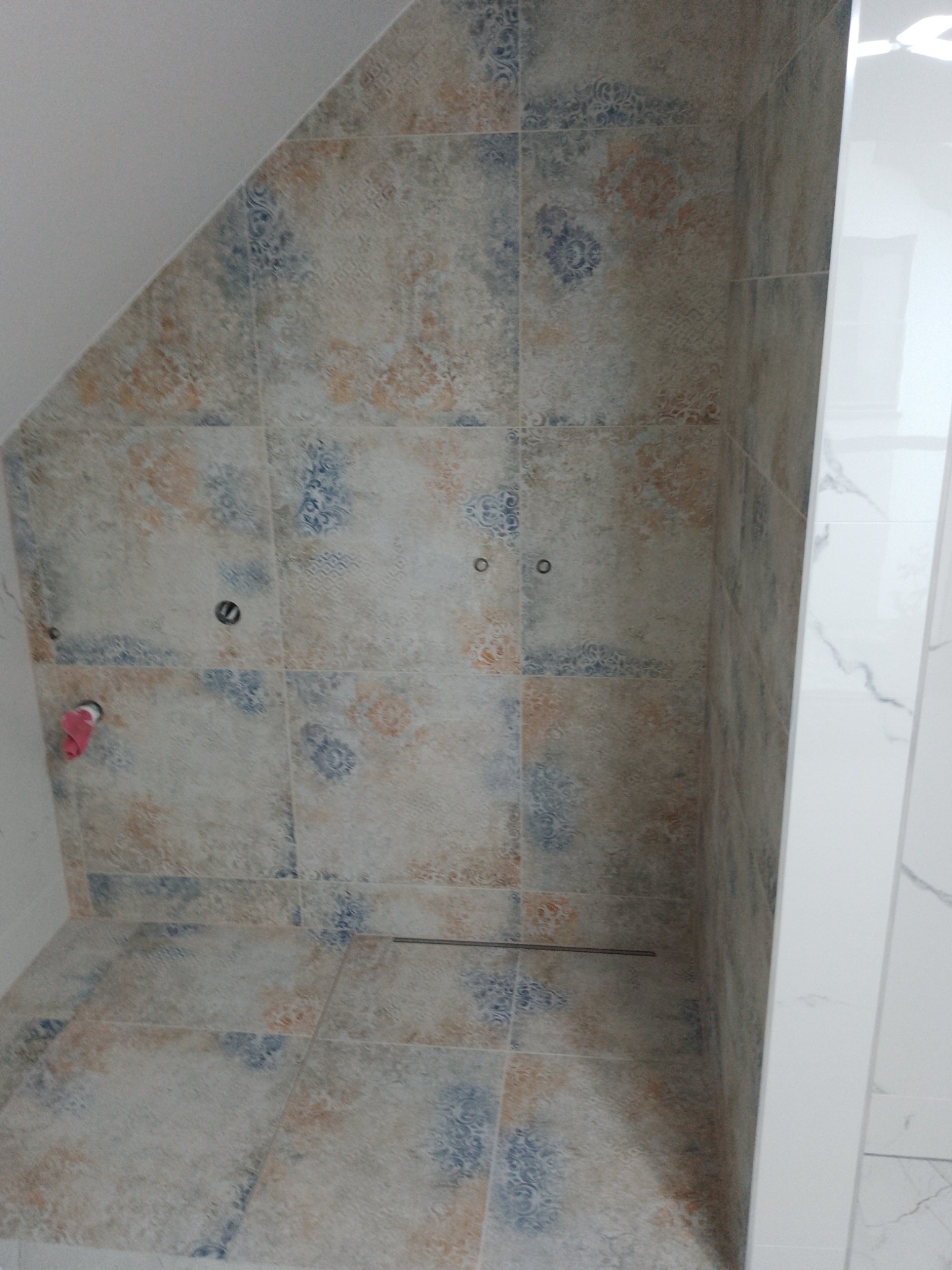 Wykończona kabina prysznicowa z dekoracyjnymi, wzorzystymi płytkami w odcieniach beżu, błękitu i brązu, z odpływem liniowym w podłodze i widocznymi przyłączami wodnymi na ścianie.