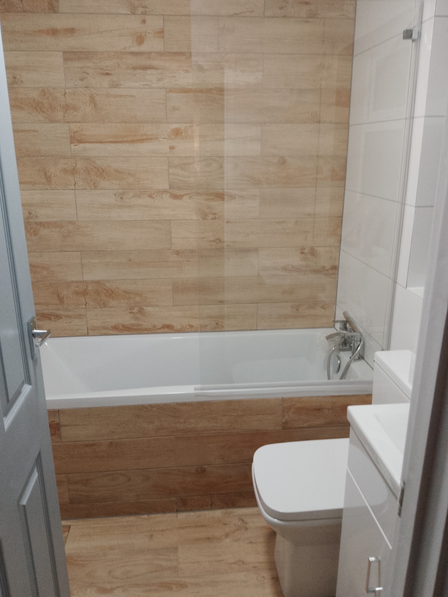 Wykończona łazienka z wanną obudowaną płytkami imitującymi drewno, białą armaturą i częściowo przeszkloną ścianką prysznicową, widok zza uchylonych drzwi.