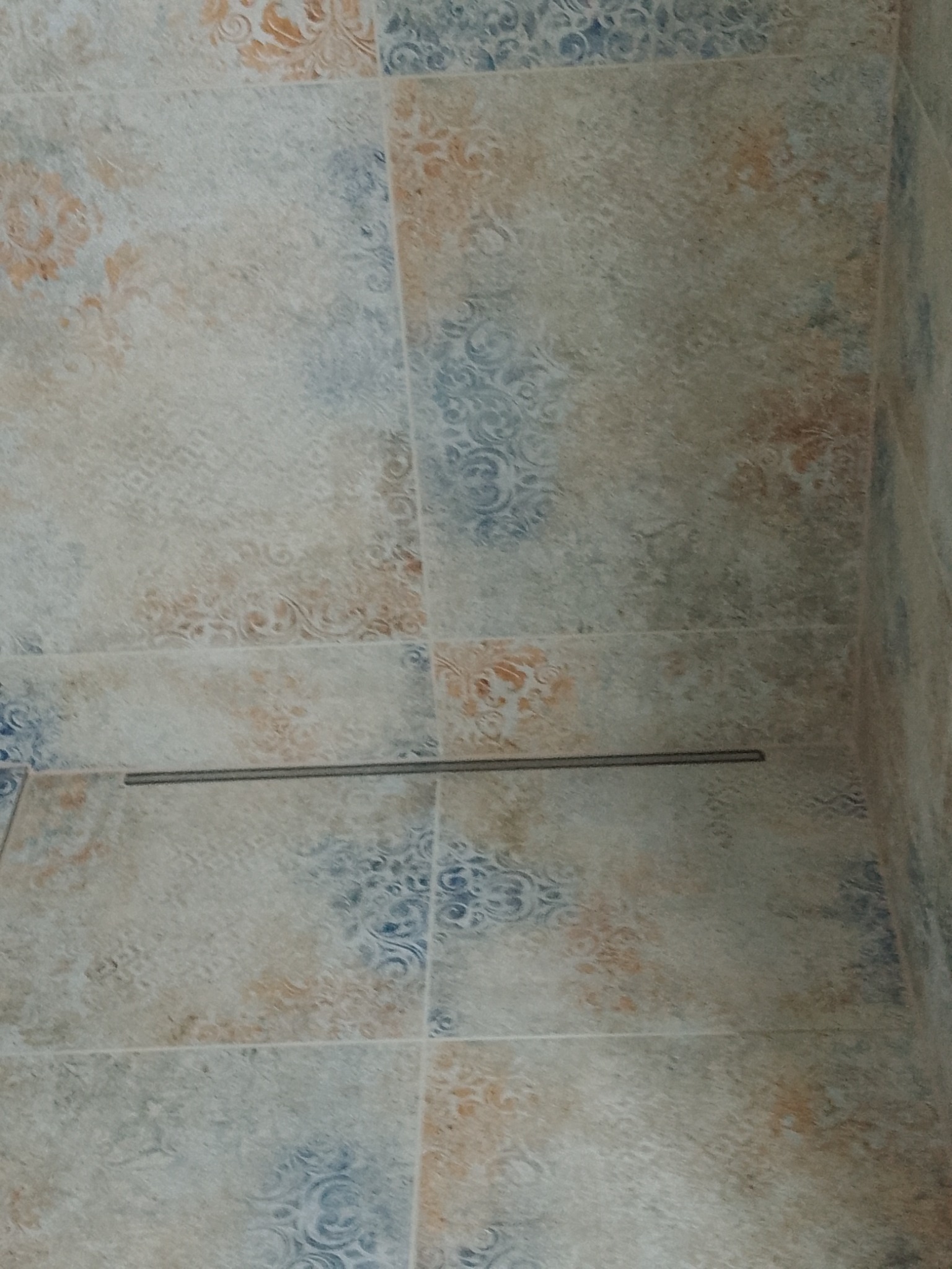 Wzorzyste, beżowo-błękitne płytki ceramiczne na ścianie, z widoczną metalową listwą dekoracyjną.