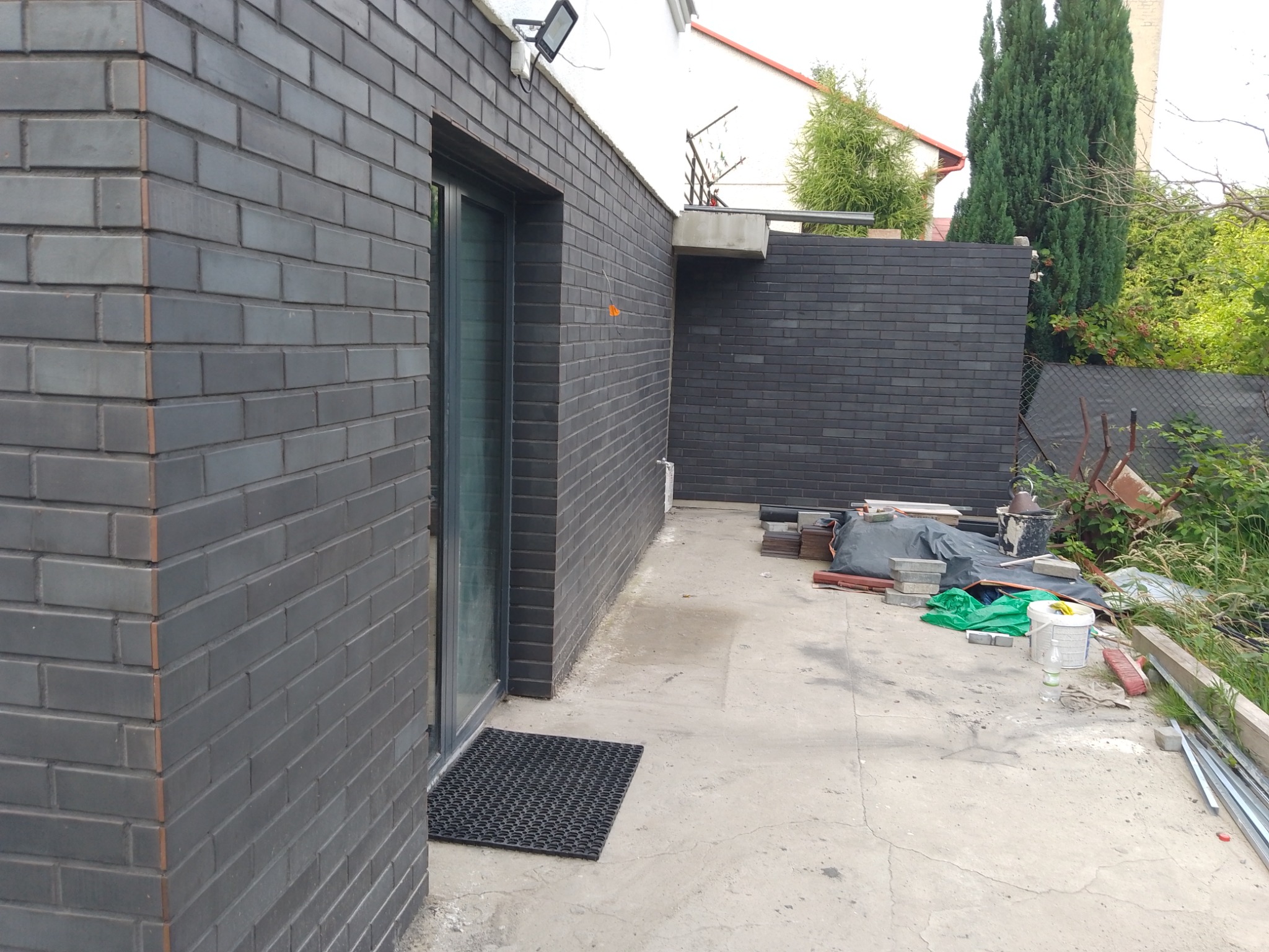 Elewacja budynku wykończona ciemnoszarą cegłą klinkierową, widoczny fragment ściany z oknem i czarną wycieraczką, w tle materiały budowlane i elementy ogrodu.
