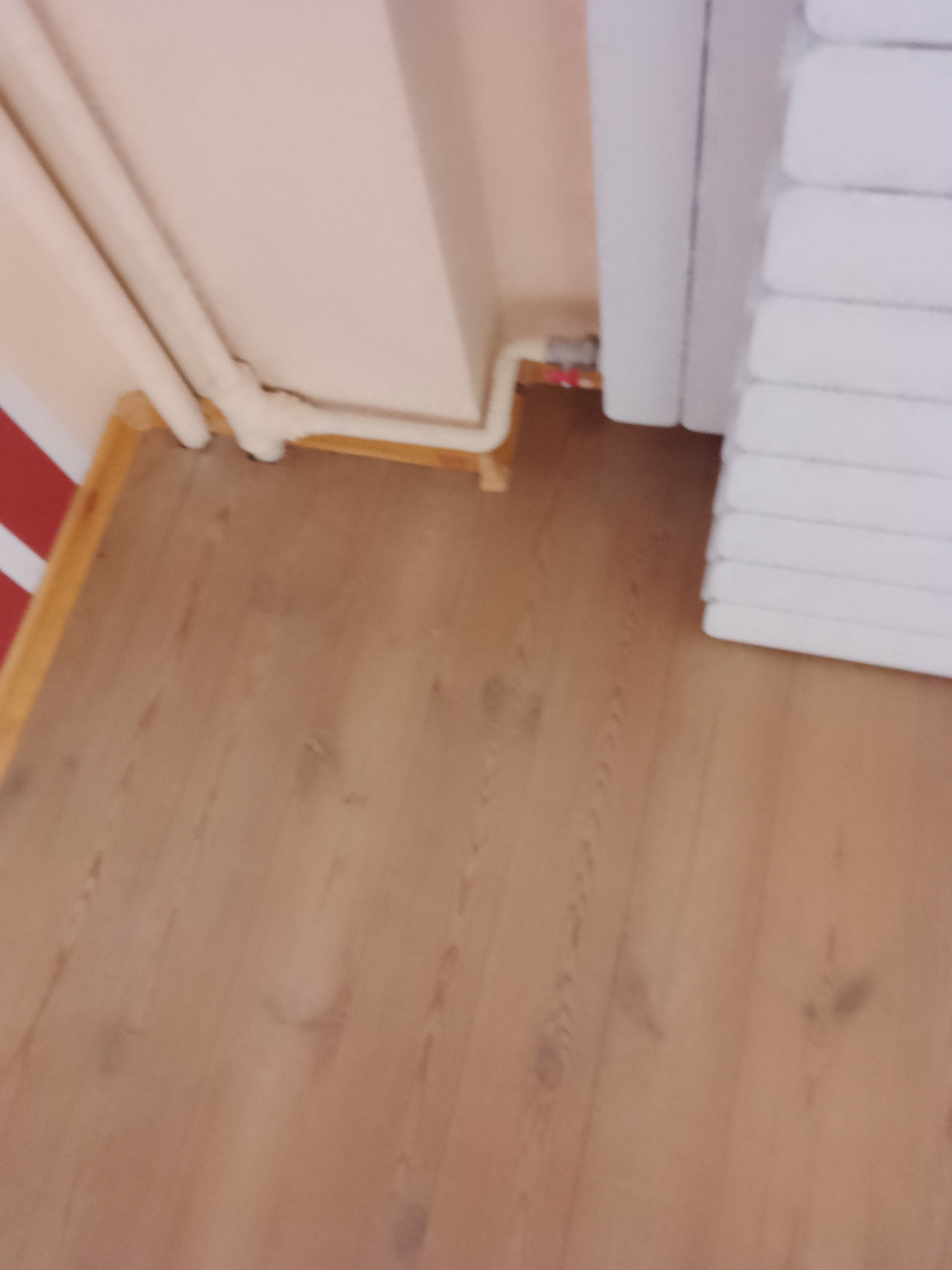 Jasnobrązowe panele podłogowe z widocznym fragmentem grzejnika i rury przy ścianie. Ujęcie z niskiego kąta, blisko podłogi.