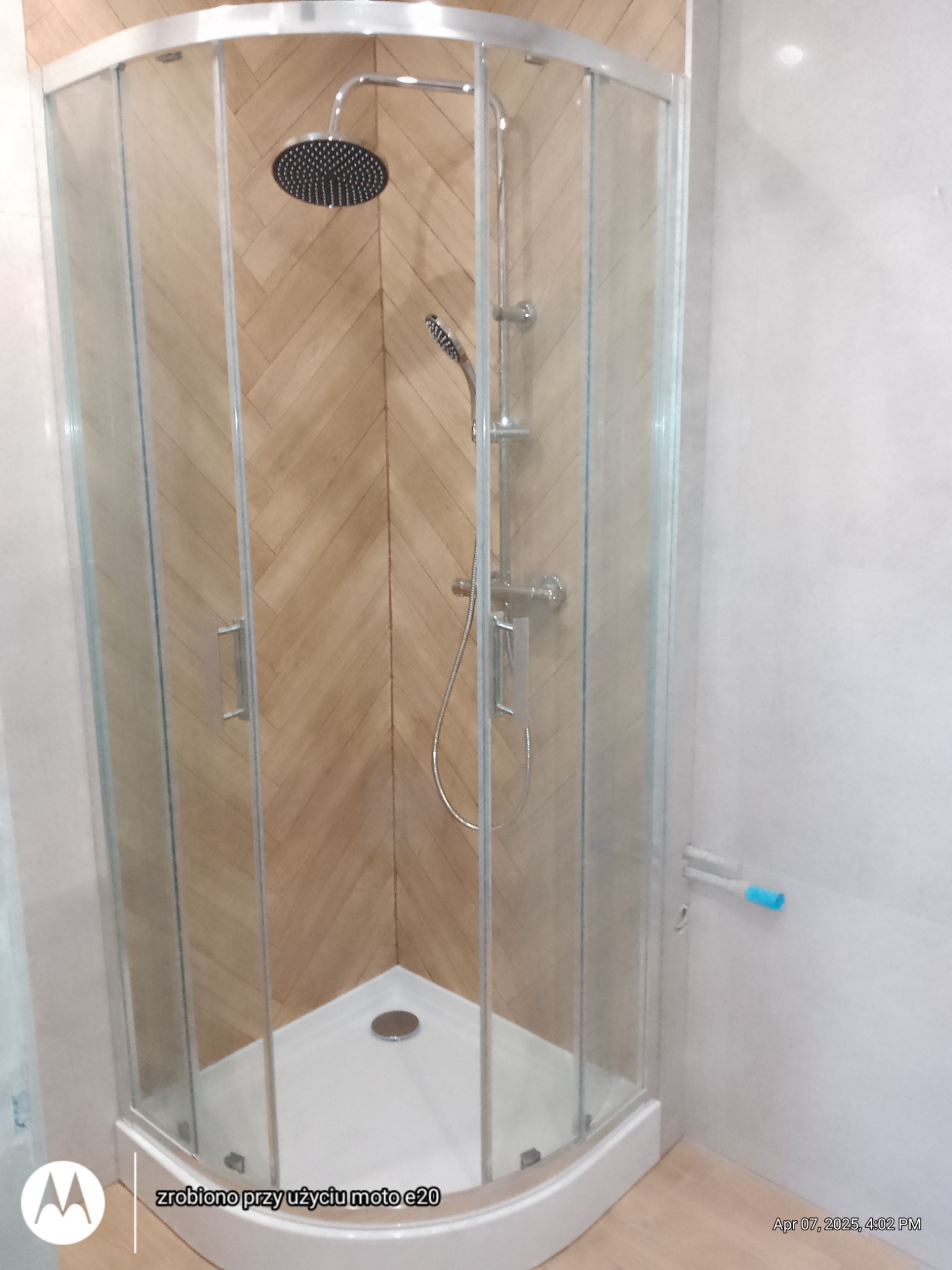 Nowoczesna kabina prysznicowa z deszczownicą i wykończeniem ścian w jodełkę. Biały brodzik i chromowane elementy armatury. Jasne, minimalistyczne wnętrze łazienki.