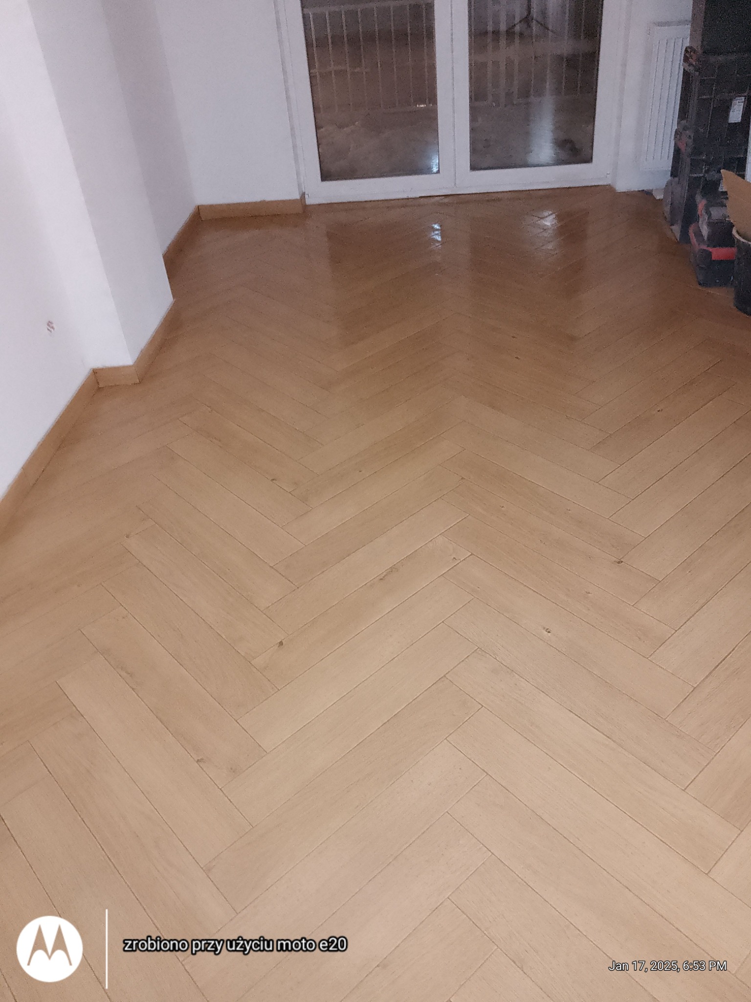 Ułożony parkiet w jodełkę francuską, wykończony listwami przypodłogowymi. Widoczne drzwi balkonowe i fragment grzejnika. Parkiet świeżo po lakierowaniu, z połyskiem.