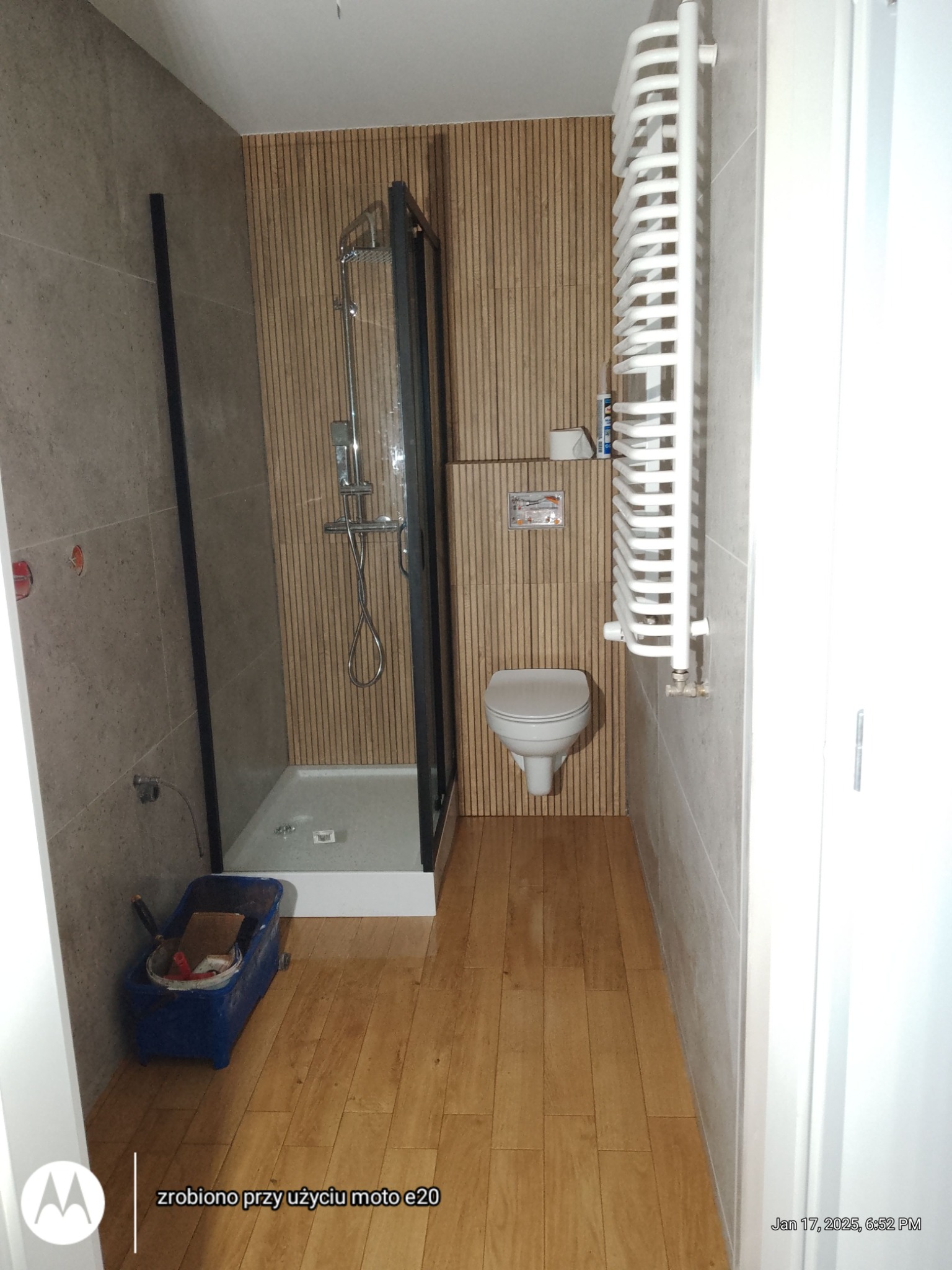 Wykończona łazienka z kabiną prysznicową, toaletą i grzejnikiem drabinkowym. Na podłodze panele imitujące drewno, ściany wyłożone płytkami w odcieniach szarości i beżu.