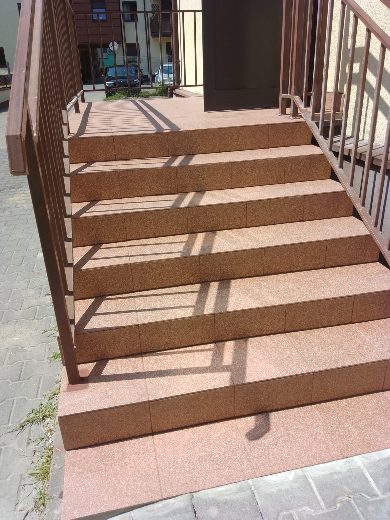 Zewnętrzne schody wejściowe wyłożone brązowymi płytkami, z metalową balustradą. Widoczne cienie rzucane przez balustradę na płytki. Tło stanowi budynek.