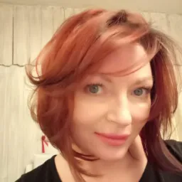 Portret kobiety z krótkimi, rudymi włosami, delikatnym makijażem i jasnymi oczami, na tle jasnej ściany z pionowymi paskami.