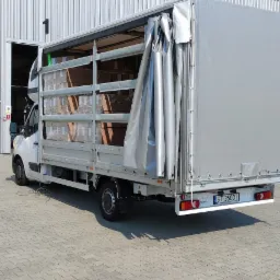 Ciężarówka dostawcza z otwartą plandeką, częściowo załadowana kartonami, stoi na brukowanym placu w Tychach, przed magazynem z blachy falistej.