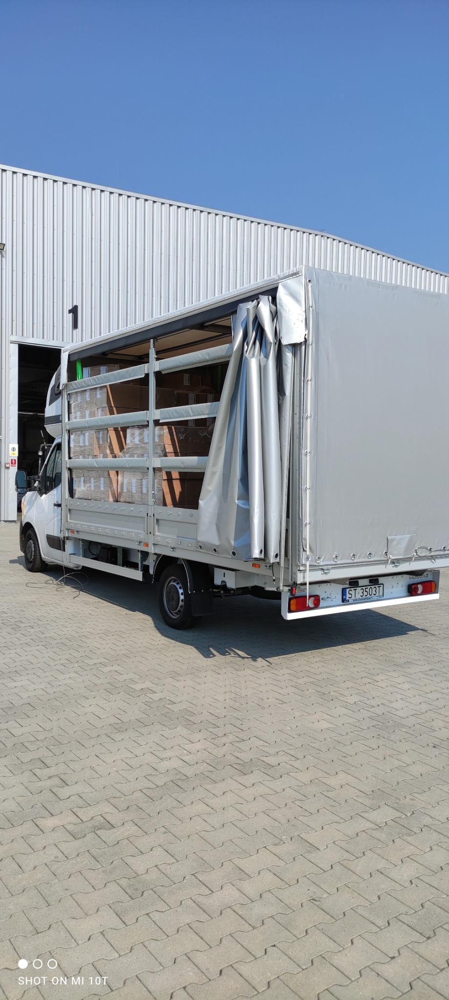 Ciężarówka dostawcza z otwartą plandeką, częściowo załadowana kartonami, stoi na brukowanym placu w Tychach, przed magazynem z blachy falistej.