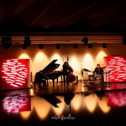 Zespół muzyczny grający jazz na scenie z czerwonymi ekranami LED w tle, reflektory punktowe oświetlają muzyków.