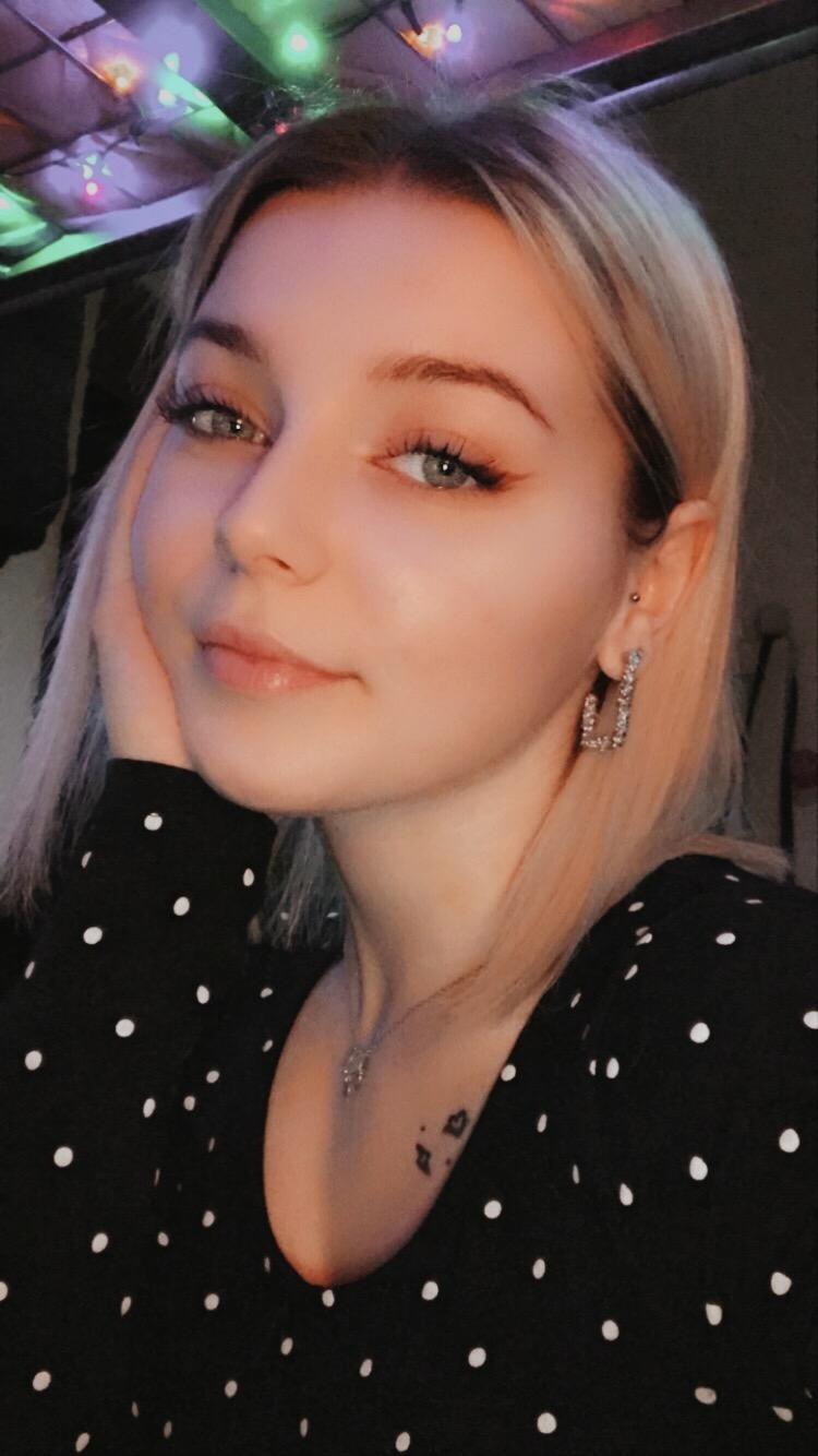 Portret młodej kobiety z blond włosami, w czarnej bluzce w białe kropki, z błyszczącymi kolczykami i delikatnym tatuażem na obojczyku, pozującej na tle nastrojowych światełek.