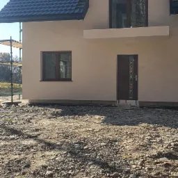 Nowoczesny dom jednorodzinny z elewacją w jasnym odcieniu, ciemną dach&oacute;wką i stolarką okienną w kolorze drewna, widoczne rusztowania z boku budynku.