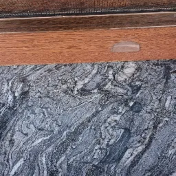 Szczegół okna z moskitierą, brązową ramą i parapetem z szarego marmuru z wyraźnym wzorem żyłkowania na tle jasnej ściany.