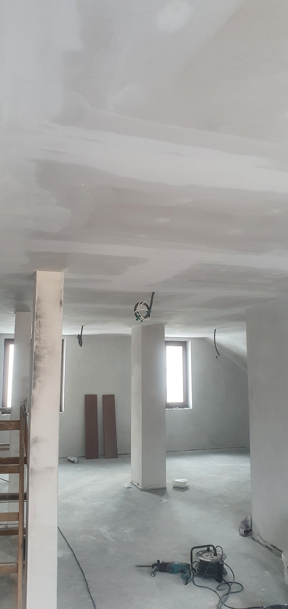 Wnętrze w trakcie prac wykończeniowych: sufit i ściany po szpachlowaniu, widoczne narzędzia (wiertarka udarowa, przedłużacz) na podłodze, wystające przewody elektryczne, drewniane płyty oparte...