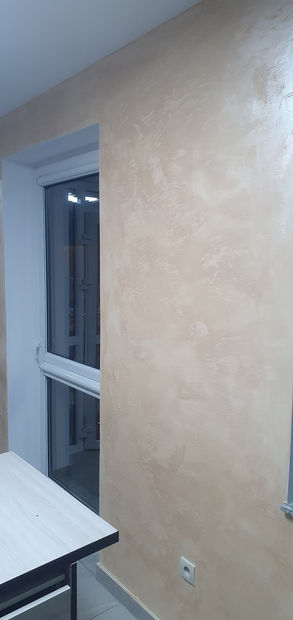 Ściana w pokoju po malowaniu z efektem dekoracyjnym w odcieniach beżu i kremu, widoczny fragment okna z roletą i gniazdko elektryczne.