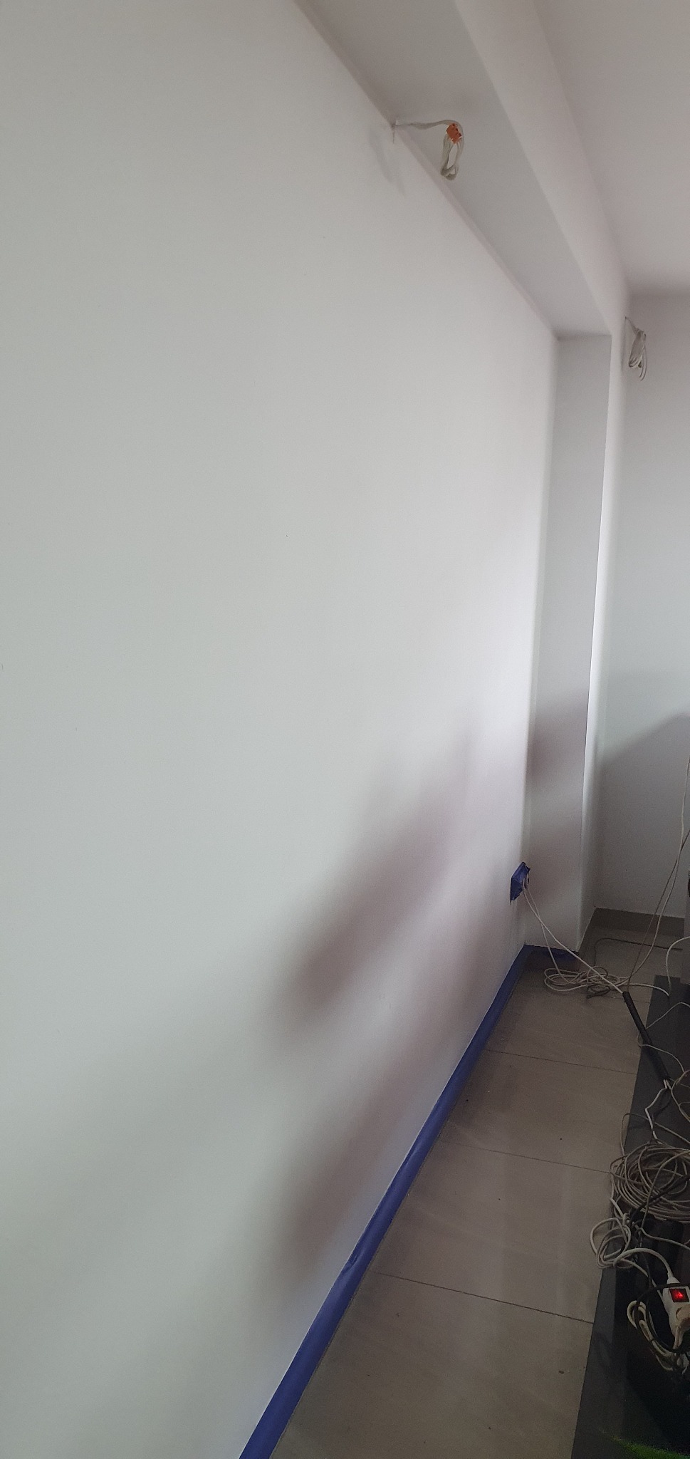 Świeżo pomalowana, biała ściana z niebieską taśmą malarską przy podłodze, widoczne gniazdko elektryczne i kable na podłodze.