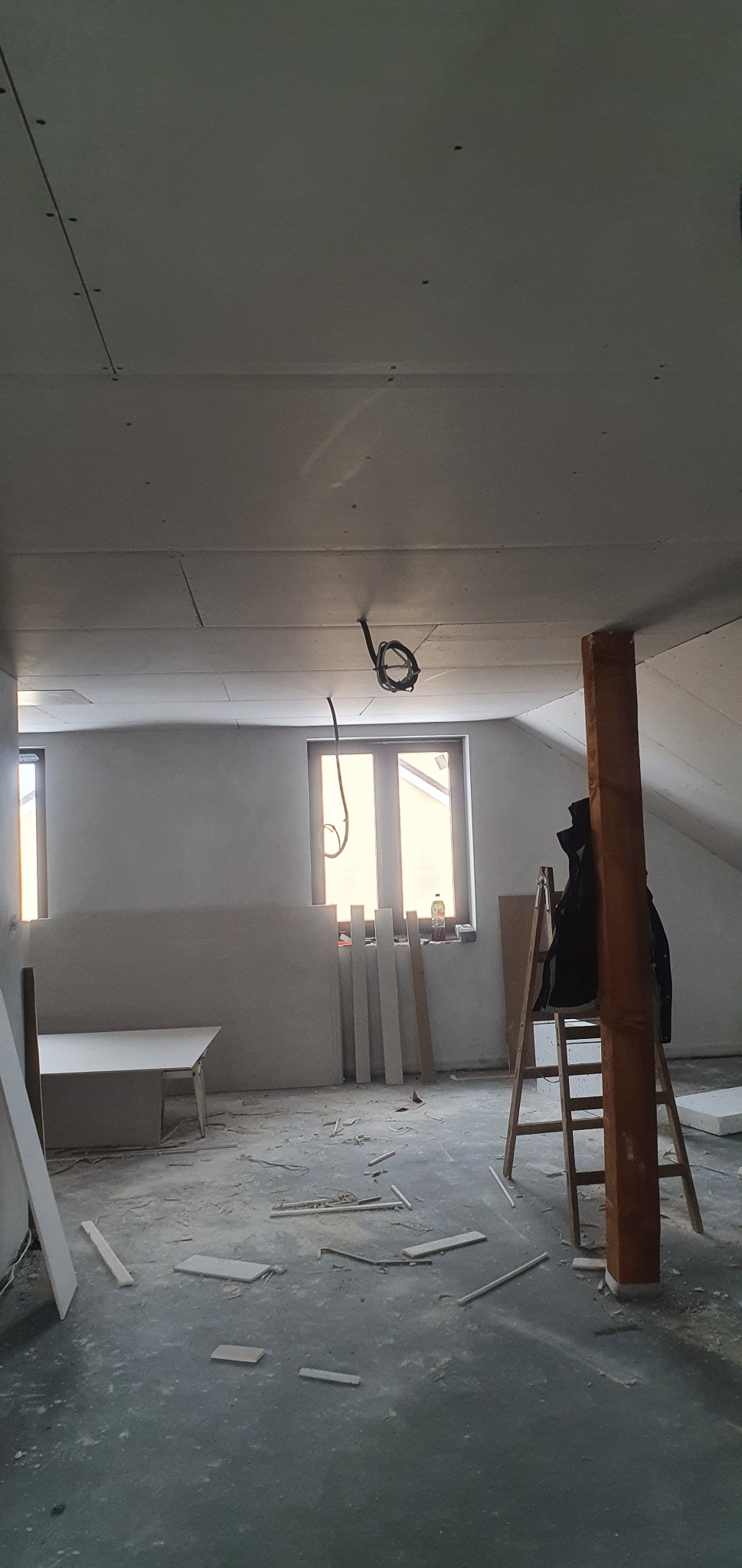 Poddasze w trakcie prac wykończeniowych: zamontowane płyty gipsowo-kartonowe na suficie, widoczne kable, drabina oparta o drewniany słup, resztki materiałów budowlanych na podłodze.