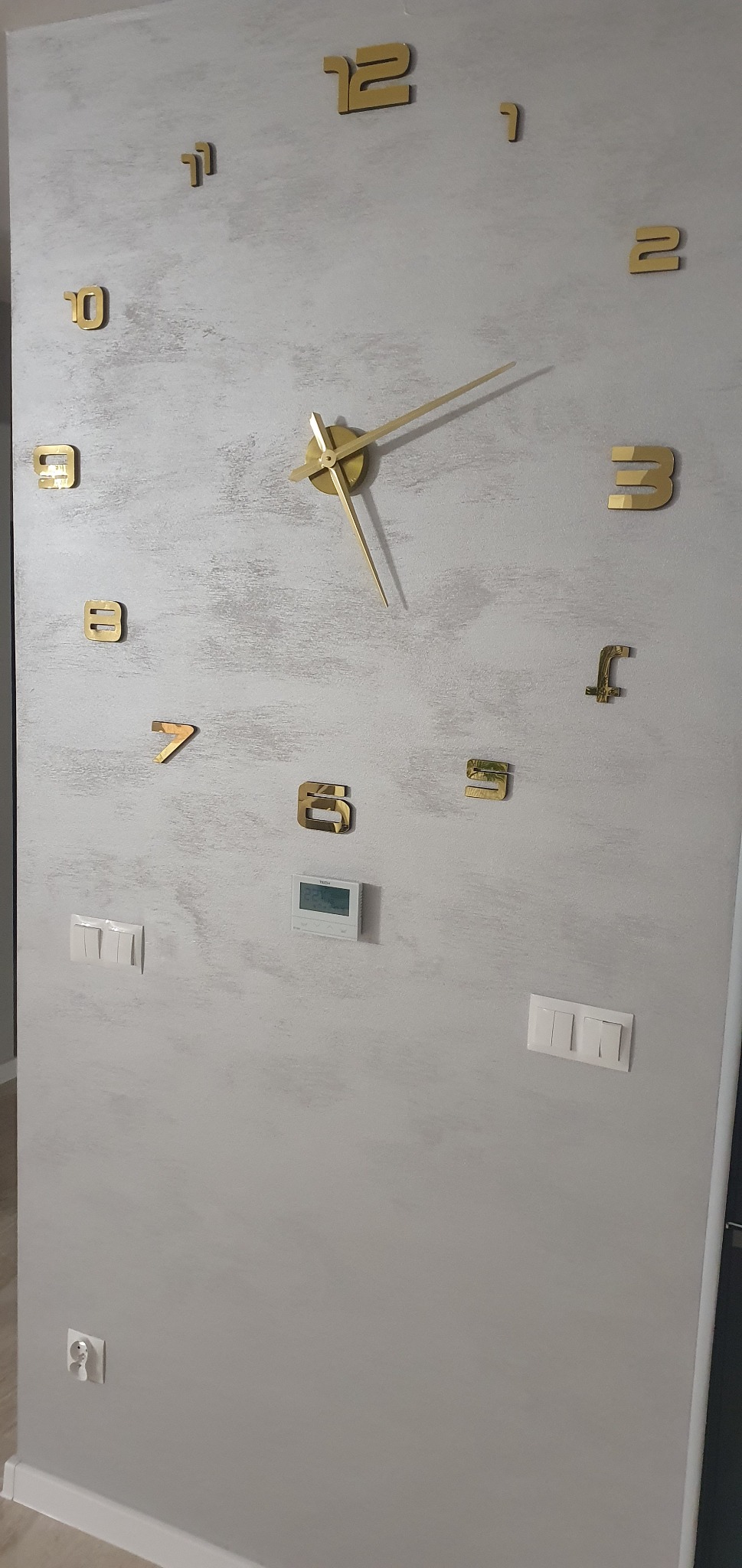 Szara ściana z efektem dekoracyjnym, na której umieszczono złote, przestrzenne cyfry tworzące duży zegar ścienny; widoczne także białe włączniki światła i termostat.