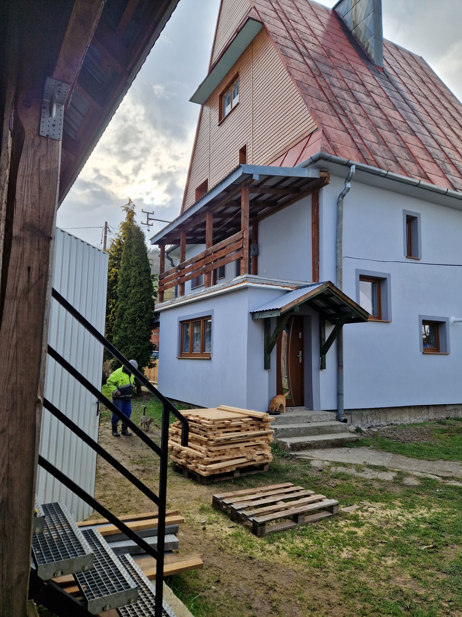 Dwukondygnacyjny dom z balkonem w Brunarach, z drewnianą konstrukcją i szarą elewacją, widziany z perspektywy schodów, z deskami przygotowanymi do montażu i kotem przy wejściu.